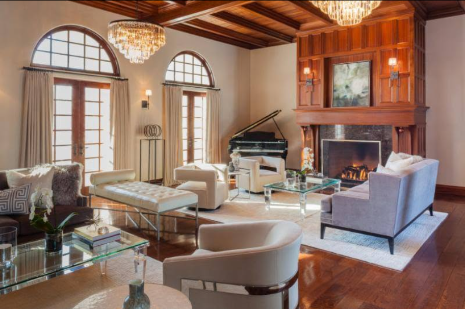 Elegant living room with a grand piano near a stone fireplace, chandelier, and white sofas arranged around glass coffee tables